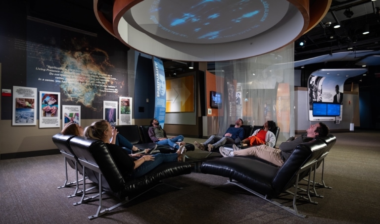 Six museum visitors in an exhibit sitting in reclined chairs looking up at a projection on the ceiling