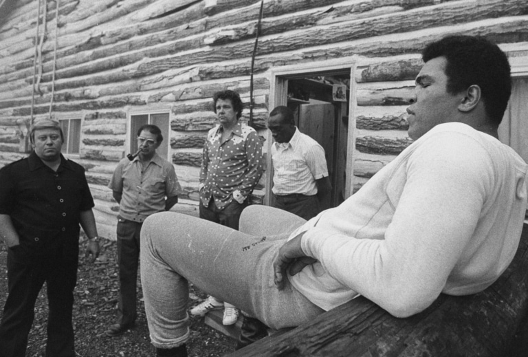 Black and white photo of Muhammad Ali sitting on wooden logs outside Deer Lake as his friends gather nearby