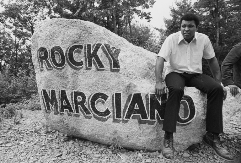 Black and white photo of Muhammad Ali sitting on large rock with painted on words "Rocky Marciano"