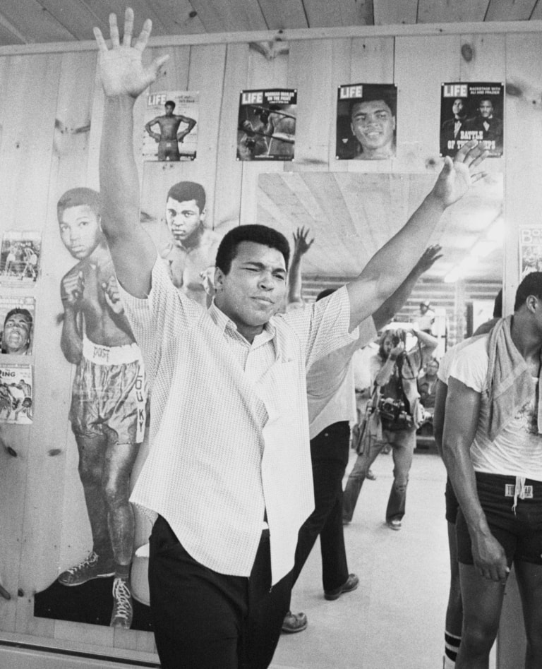 Black and white photo of Muhammad Ali raising both his hands in the air as a photographer is taking his picture