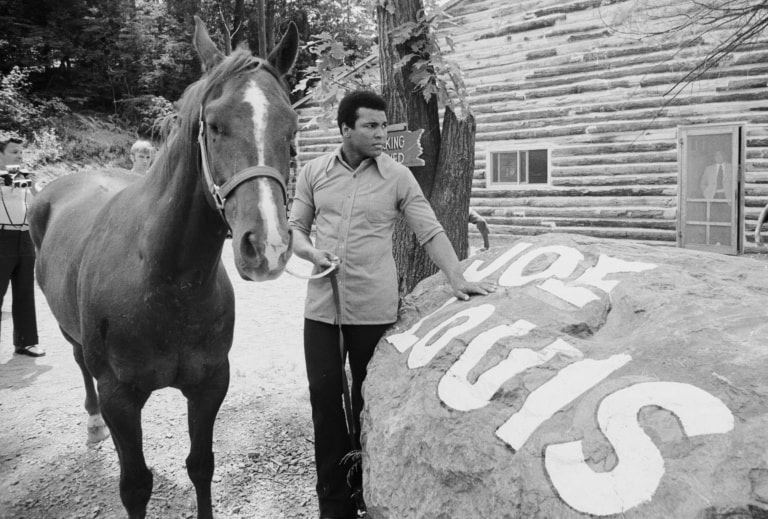 Black and white photo of Muhammad Ali leading a horse outside Deer Lake near rock with Joe Louis' name painted on top