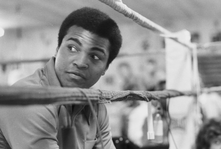 Black and white photo of Muhammad Ali peeking through ropes outside of boxing ring