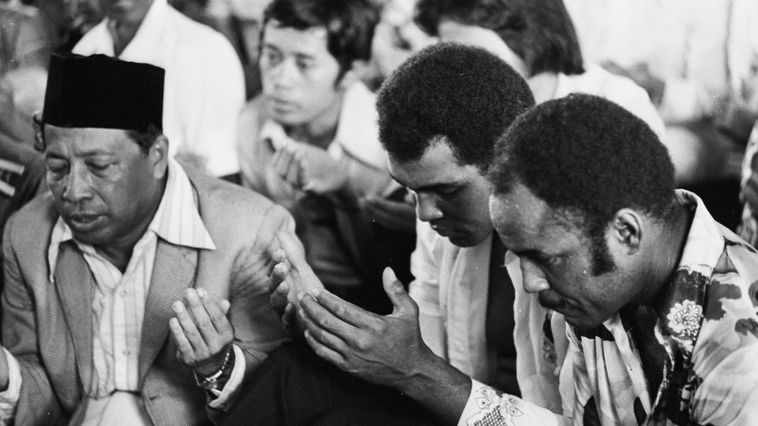 Black and white photo of Muhammad Ali in a room full of men praying