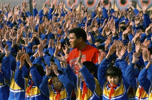 Ali in Beijing in a red sweatshirt, surrounded by children in blue and yellow uniforms with their arms raised