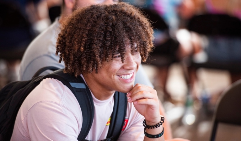 A student wearing a t-shirt and a backpack sits, listens, and smiles