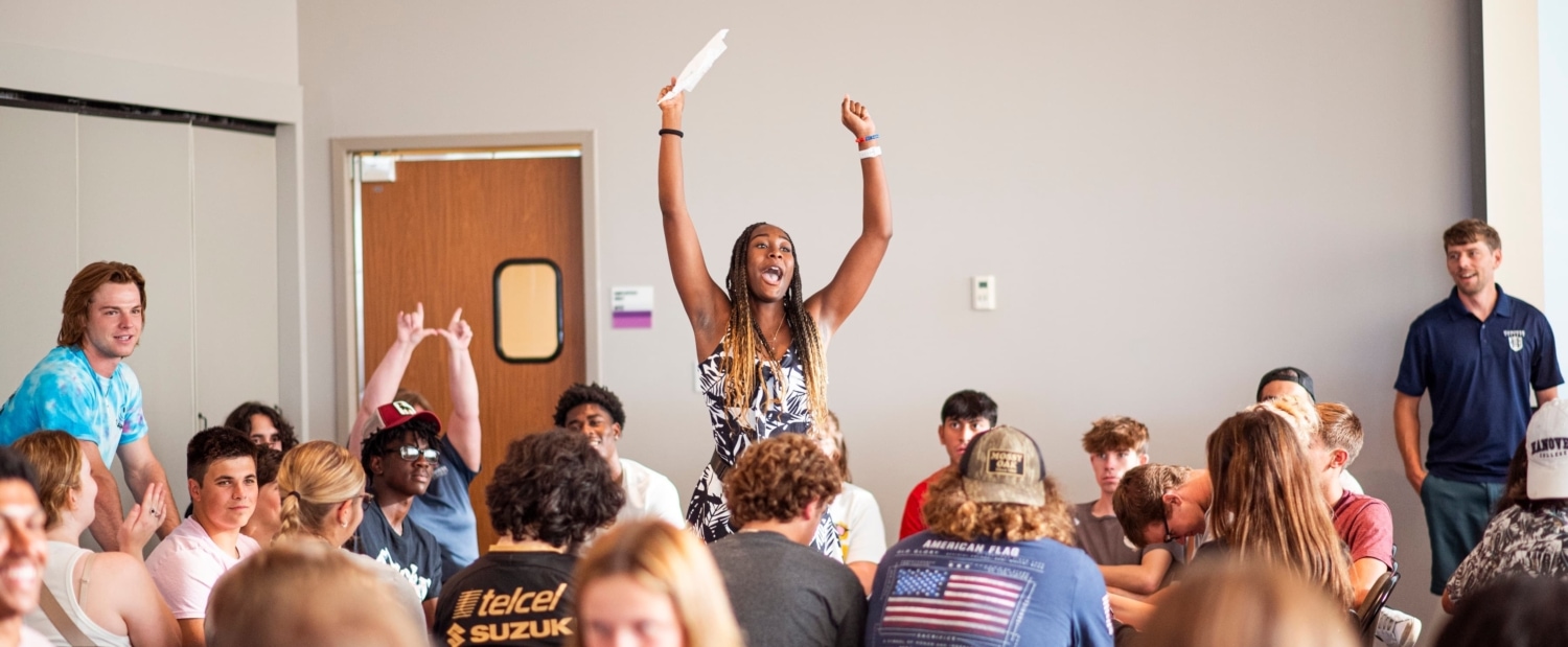 Woman standing with her arms raised high amidst a room full of college students