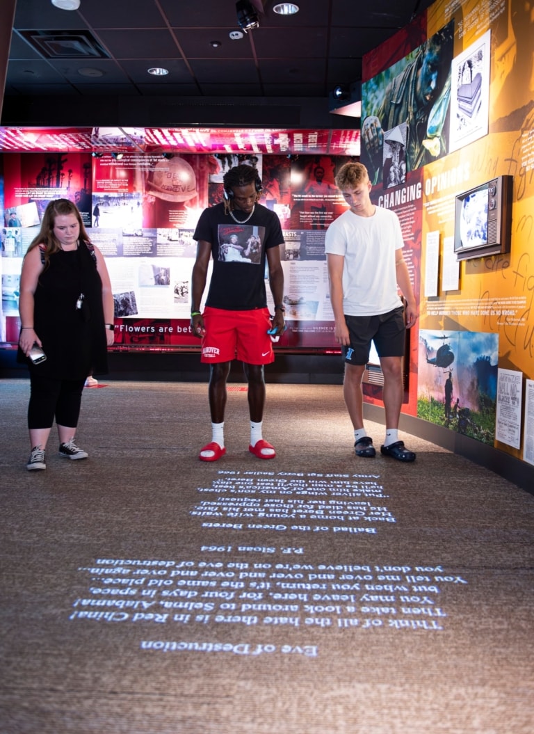 Three college students stand in a gallery at the Ali Center reading text projected onto the floor