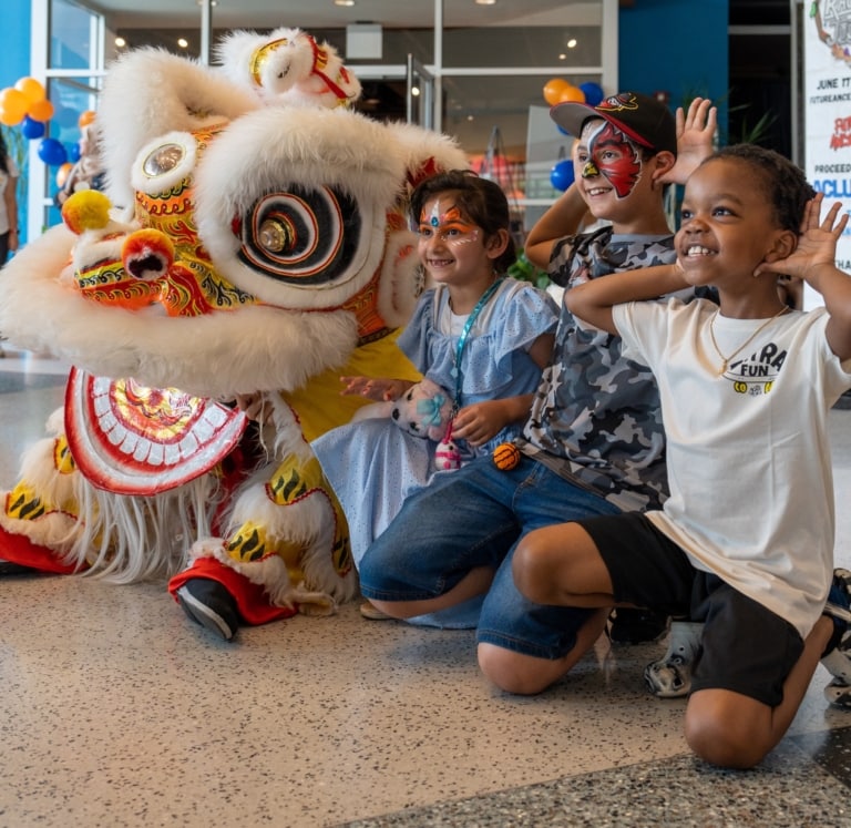 Three kids, two with face paint, pose for a photo next to a Chinese dragon performer in the lobby of the Muhammad Ali Center