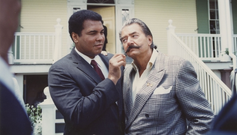 Muhammad Ali and another man with a grand mustache in suits in front of a light yellow house