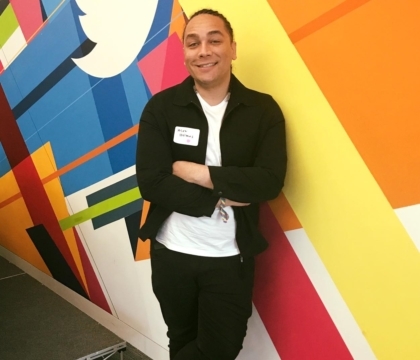 Man wearing white shirt, black jacket, black pants and sneakers stands in front of colorful wall with Twitter logo