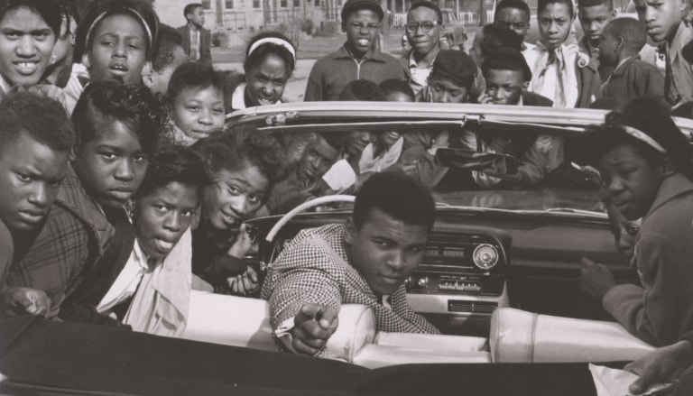 Black and white photo of Muhammad Ali as a young man sitting in a convertible and dramatically pointing while surrounded by a crowd of people around the car