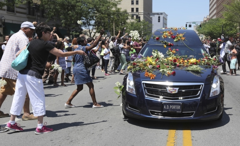 Hearse covered in flowers drives down street with crowd throwing flowers onto the car