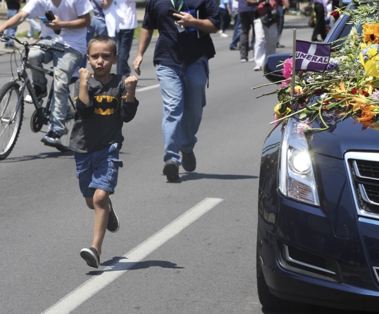 A young child with two fists in the air and a Batman t-shirt walks alongside Muhammad Ali's hearse