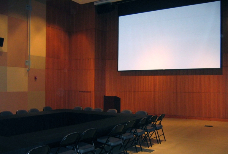 Large room with a projection screen and long black banquet tables configured in a large rectangle with folding chairs
