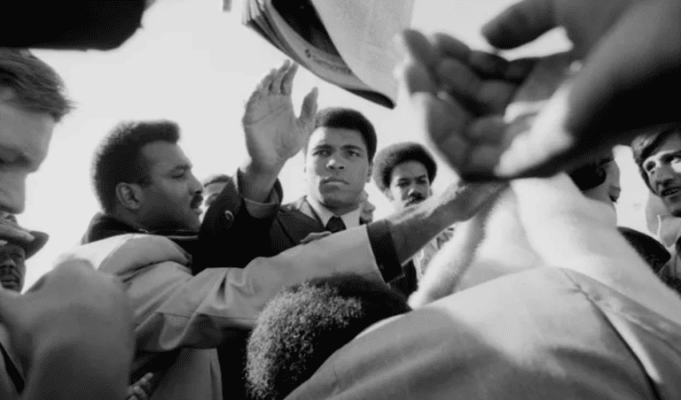 Muhammad Ali in a suit in the middle of a crowd of people reaching towards the camera