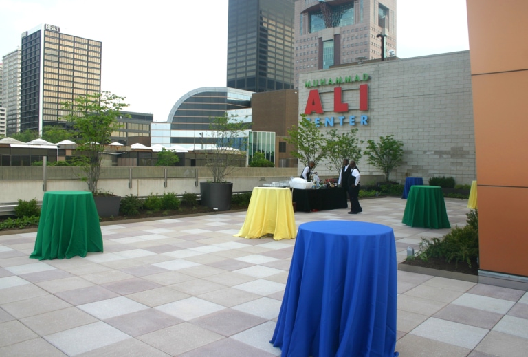 Tall round tables with colored tablecloths set up on a patio with an event bar and three event staff in the background