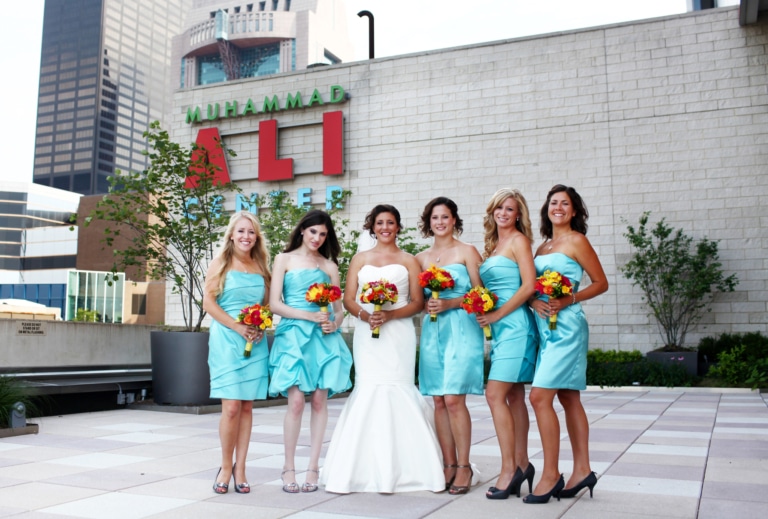 A bride and five bridesmaids in teal dresses holding bouquets and standing outside