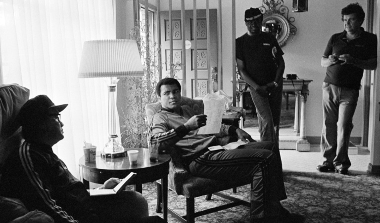 Black and white photo of Muhammad Ali sitting with friends inside a home
