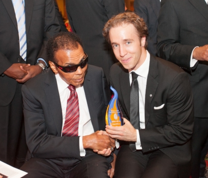 Muhammad Ali, wearing shades, shaking hands with man wearing black jacket, white shirt and black tie and holding award