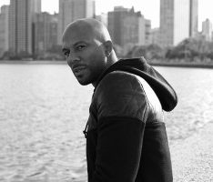 Common, wearing black hooded jacket, posing in front of body of water and city skyline