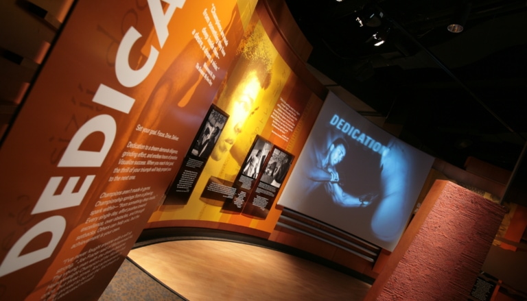 Skewed view of an exhibit gallery with the text panels on the wall and a projection screen in the distance showing Muhammad Ali boxing and the word "Dedication"