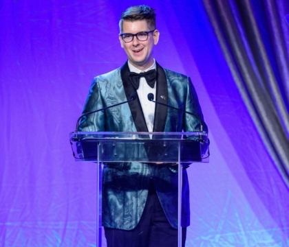 Man with glasses wearing shiny blue jacket and bowtie speaks at lecturn