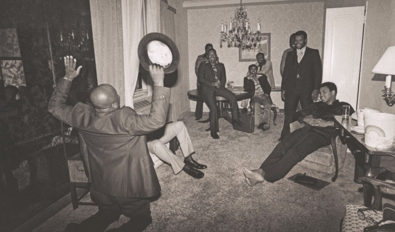 Black and white photo of Muhammad Ali in a home with several men while one man kneels on the floor and gestures wildly with his hat