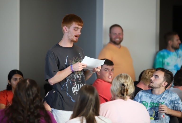 A person with red hair and facial hair stands and reads amongst a group of fellow college students