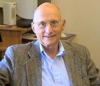 Man wearing glasses with gray suit jacket and blue shirt smiles for camera in front of office