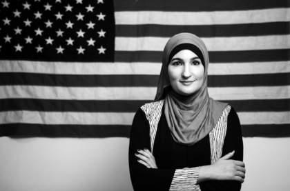Black and white portrait photo of a woman in a headscarf standing in front of an American flag