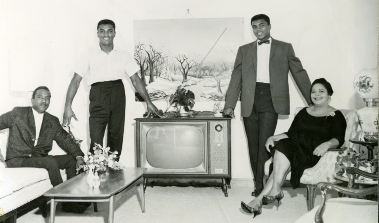 Black and white photo of Muhammad Ali as a young man in a living room with three other people
