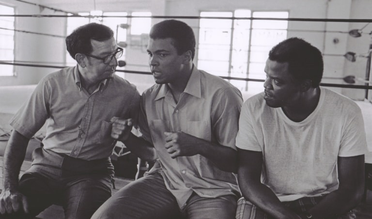 Black and white photo of Muhammad Ali and three men sitting on the edge of a practice ring talking