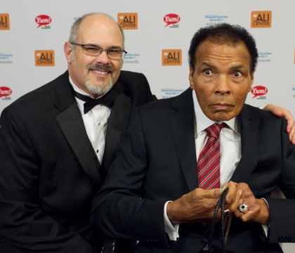 Man wearing glasses with black jacket, white shirt and bow tie posing with Muhammad Ali