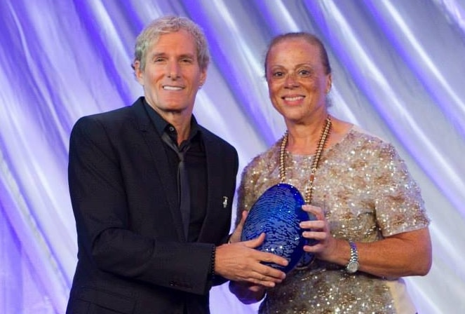 Michael Bolton, wearing black jacket, black shirt and black tie, posing with Lonnie Ali and award