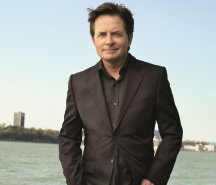 Michael J. Fox posing with black suit and black shirt in front of body of water