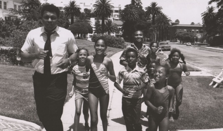 Black and white photo of Muhammad Ali with his children running outdoors on a sidewalk