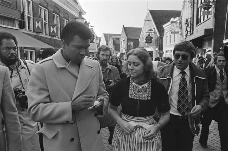 Black and white photo of Muhammad Ali in a coat, signing an autograph book for a woman in traditional Dutch dress surrounded by several photographers