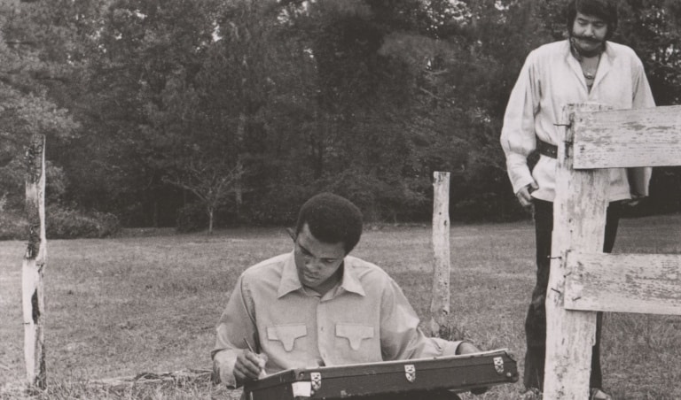 Black and white photo of Muhammad Ali as a young man, seated, drawing outdoors while another man stands nearby and looks at his work