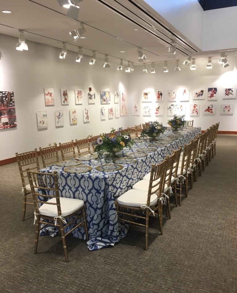 A long banquet table set with chairs, chargers, and floral arrangements in an art gallery