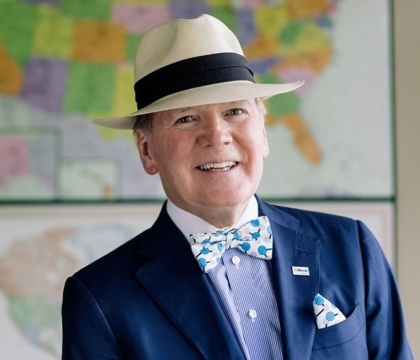 Man wearing blue jacket, blue shirt, bowtie and white hat smiles in front of world maps