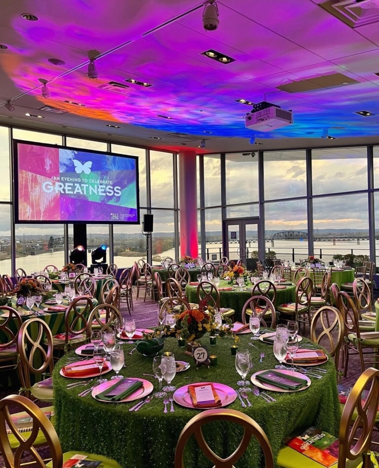 Event space with large windows, a projection screen, banquet rounds and chairs decorated for an awards event