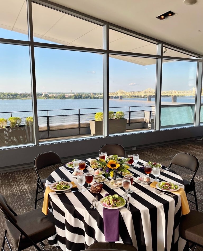 A round table with a bold stripe tablecloth, salads, water, and iced tea in front of a large window looking out onto a river and bridge