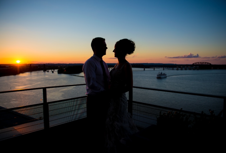 A couple standing on a balcony in front of a sunset over a river