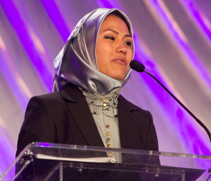 Woman wearing silver hijab and black jacket speaking at lecturn