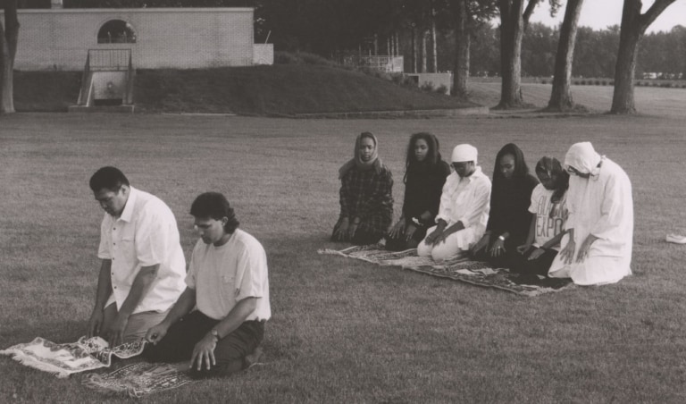 Black and white photo of Muhammad Ali with family and guests kneeling and praying on a lawn