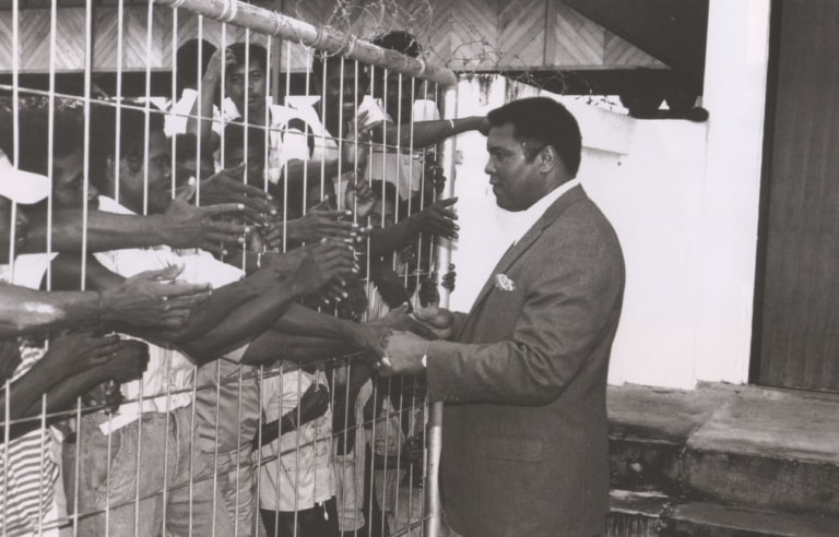 Black and white photo of Muhammad Ali in a suit shaking hands with some of the many men behind a fence and barbed wire