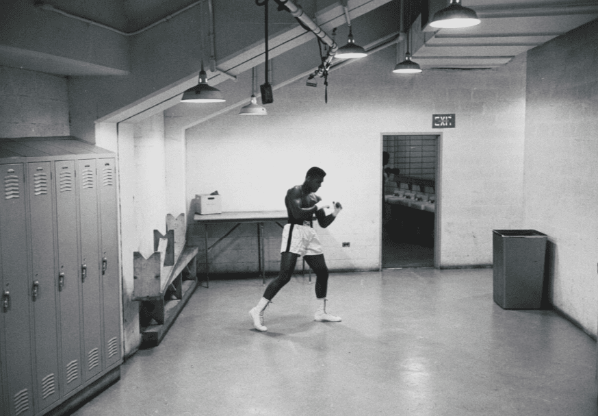 Black and white photo of Muhammad Ali in boxing gear in a locker room
