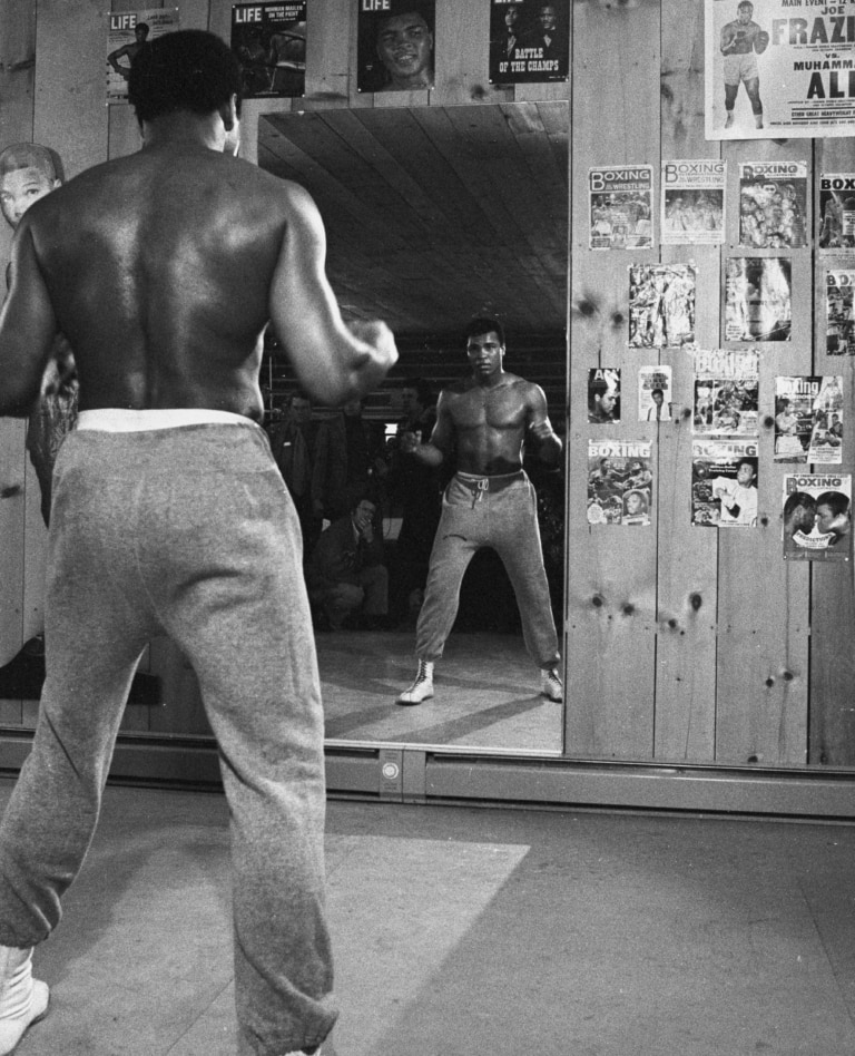 Black and white photo of Muhammad Ali looking at himself in a mirror with boxing programs taped on wall nearby