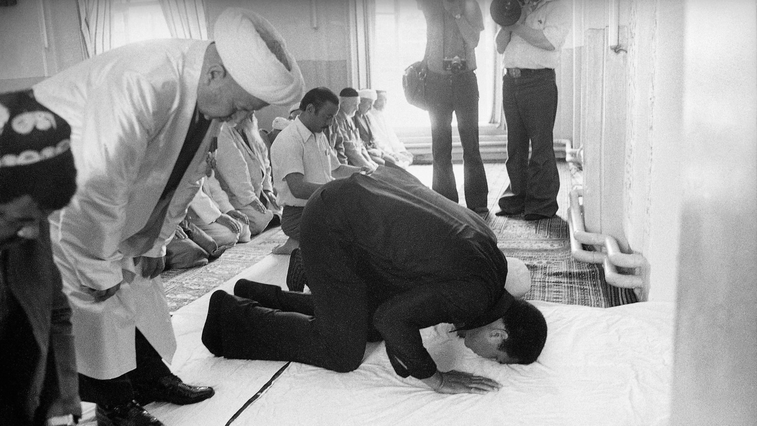 Black and white photo of Muhammad Ali praying with several other Muslim men