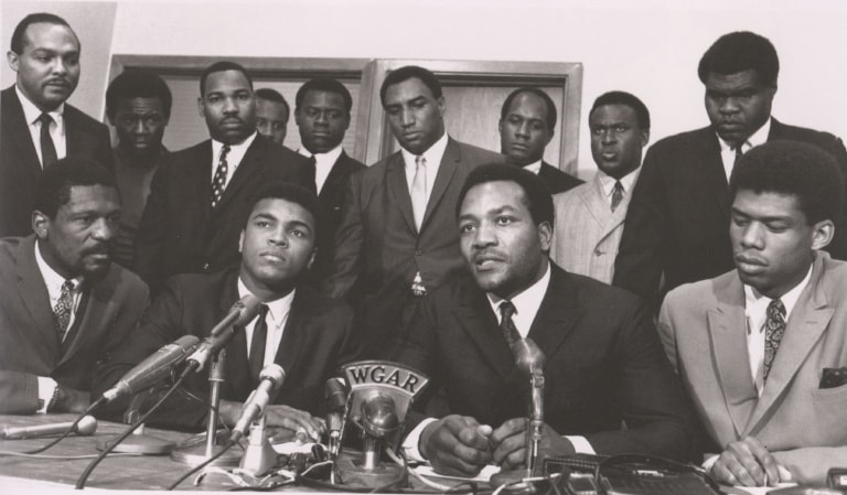 Black and white photo of Ali at the Cleveland Summit behind microphones and with a number of other men in suits
